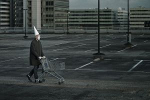 Man met clownsmake-up en puntmuts duwt winkelwagen op lege parkeerplaats met kantoorgebouwen.