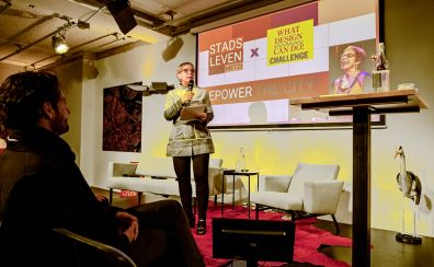 Vrouw spreekt op podium voor projectie "STADS LEVEN TRACY METZ EMPOWER THE CITY" met een lachende vrouw erop.