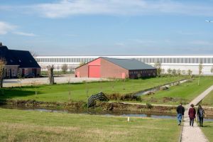 Grote, witte industriële hal achter een rode schuur en boerderij, met een watergang en wandelend paar op een zonnige dag.