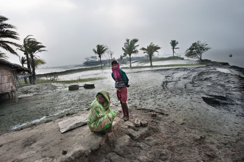 Twee vrouwen in traditionele kleding staan op een modderige oever bij palmbomen, met op de achtergrond gebouwen en water.
